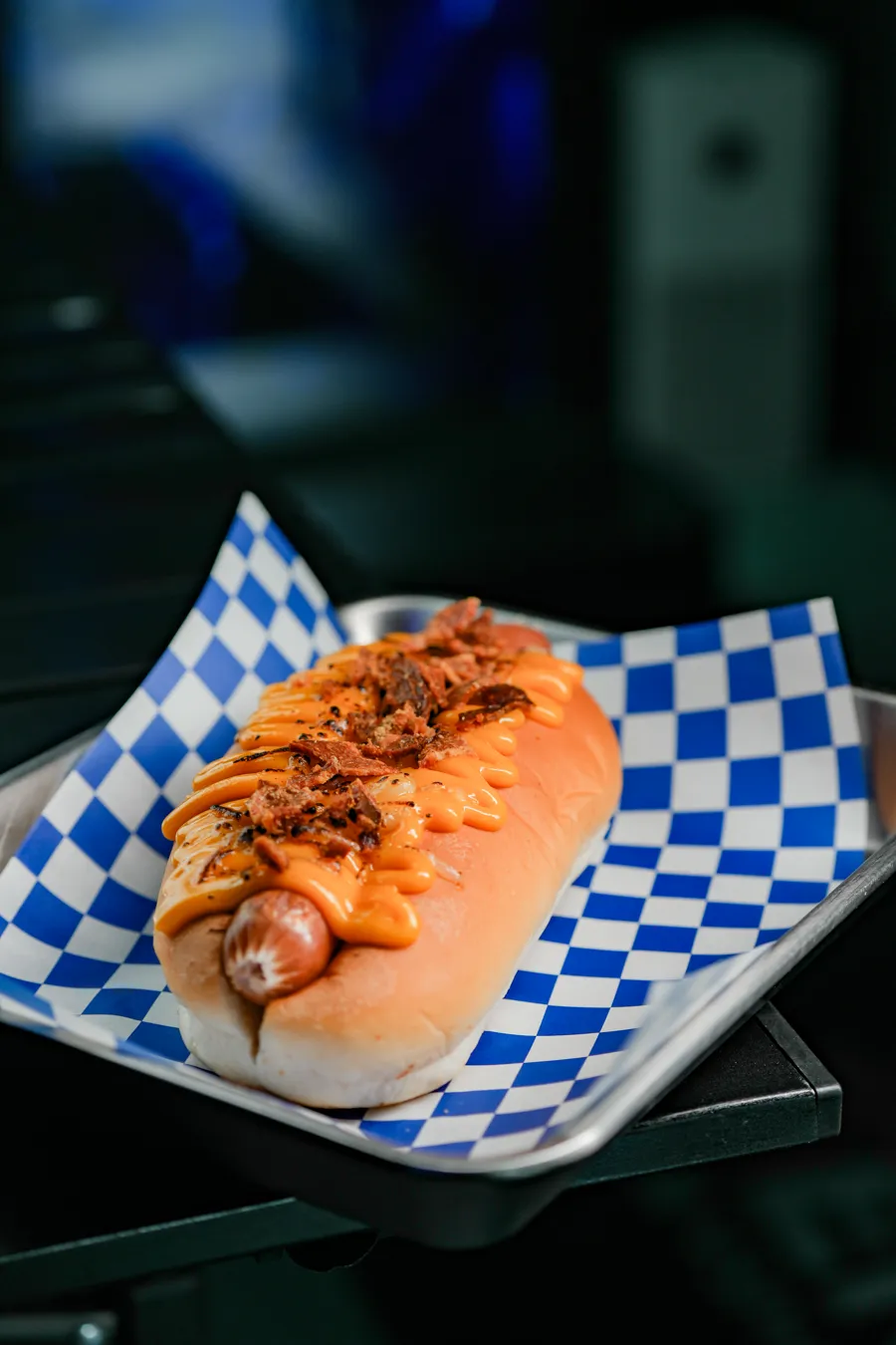 Seared bacon and cheese hot dog from the Hotdogray menu: a sausage in a bun under cheese sauce and bacon bits, on blue checkered paper