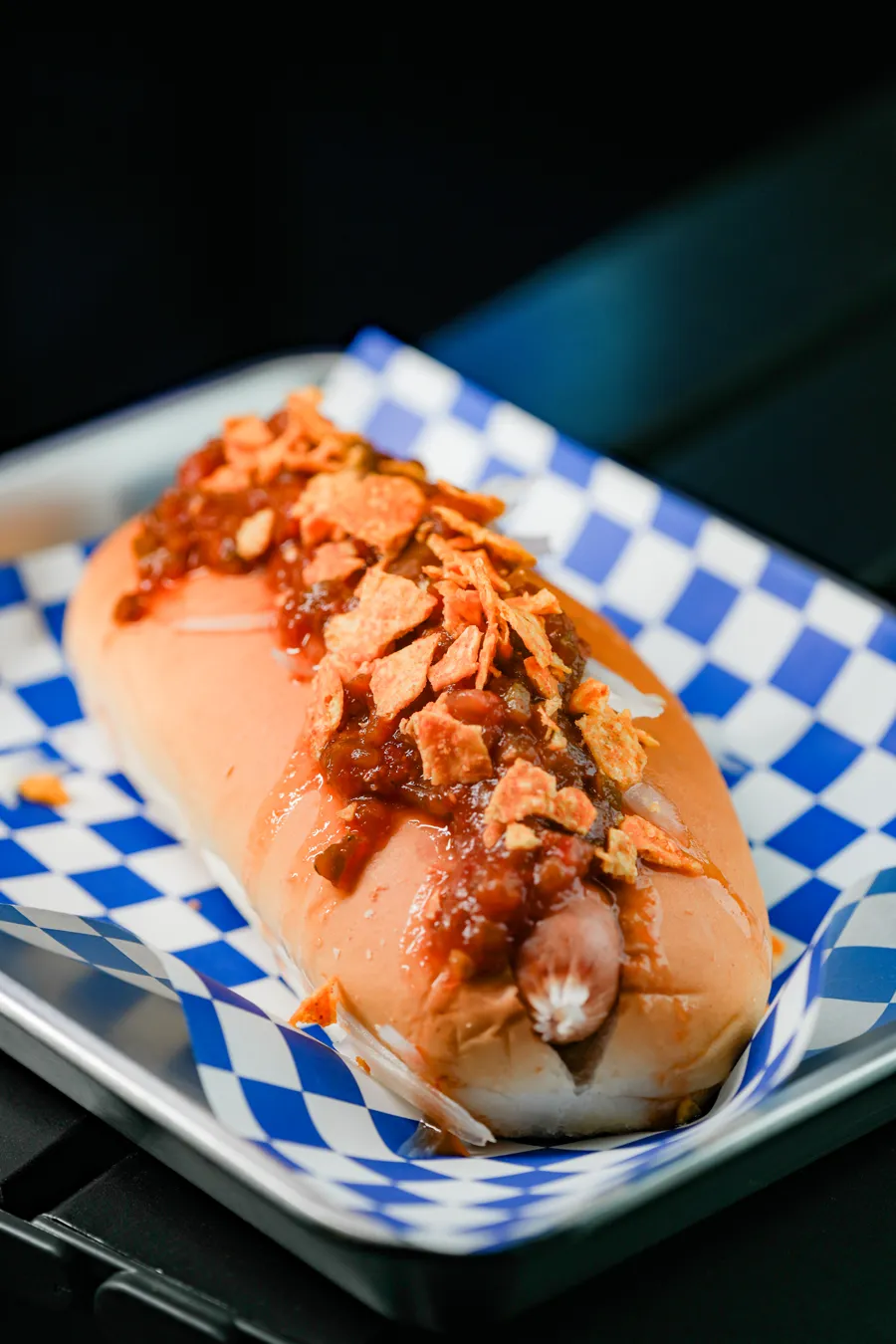 Meat sauce hot dog from the Hotdogray menu: red meat sauce over a sausage, topped with crushed chips, on blue checkered paper