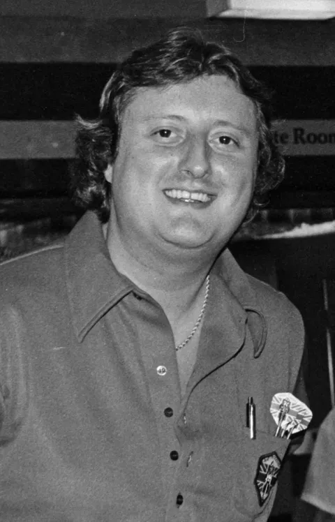 Foto en blanco y negro de 1985 de la leyenda de los dardos Eric Bristow sonriendo a la cámara con una camisa polo oscura