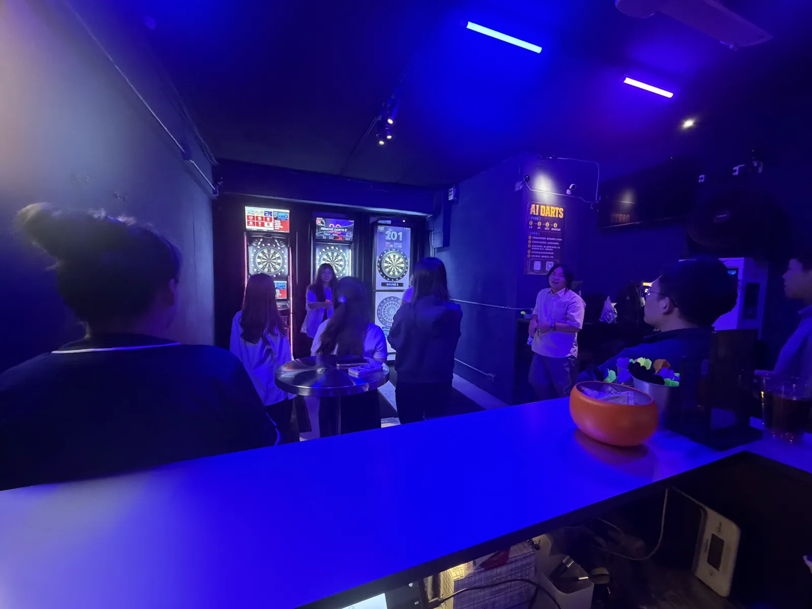 AI DARTS 店內，一群人站在飛鏢機台前輪流投鏢聊天，牆上貼著 AI DARTS 說明海報