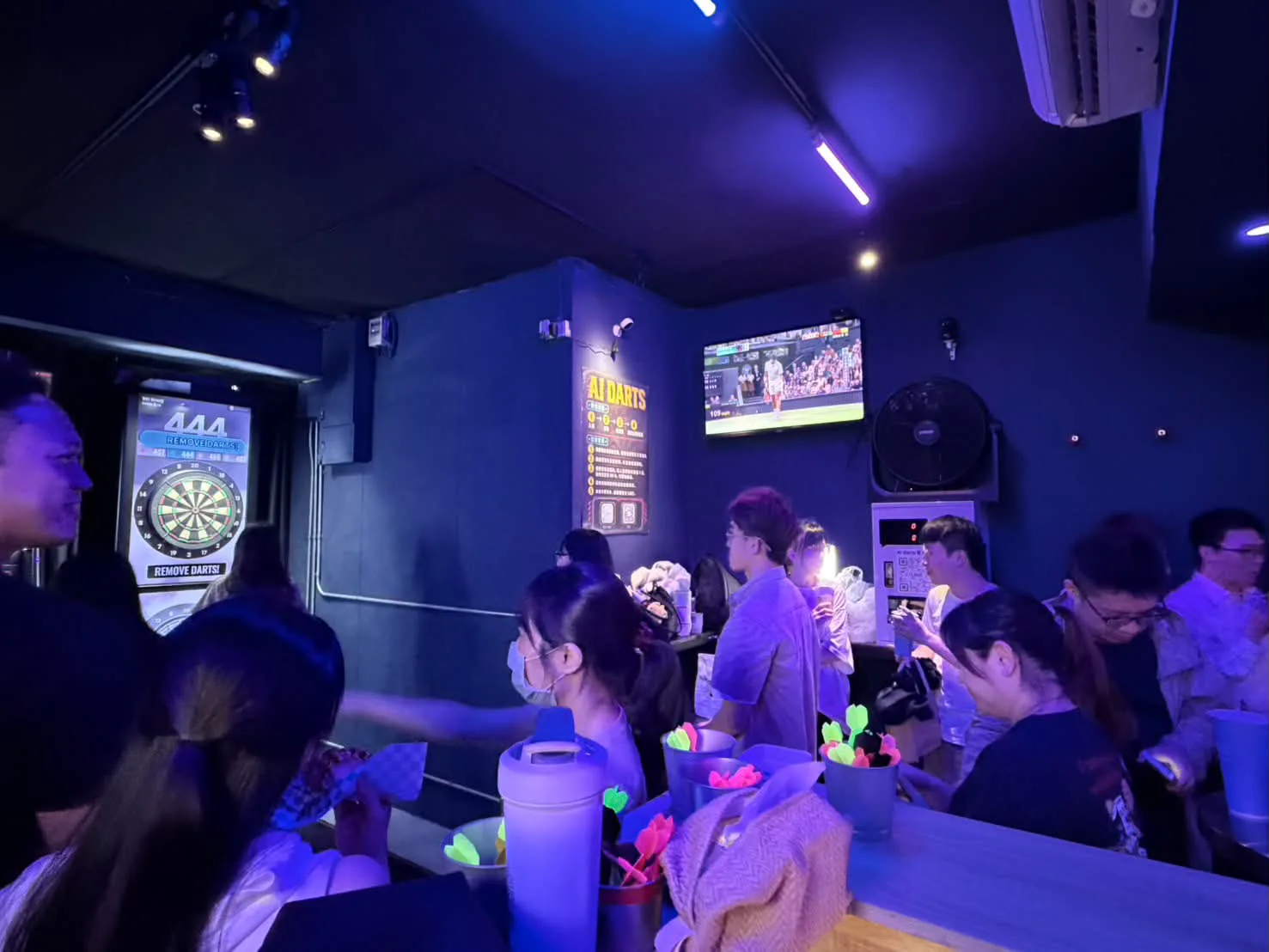 AI DARTS 吧檯前擠滿三五成群的朋友，桌上放著螢光飛鏢桶，後方電視正播著比賽
